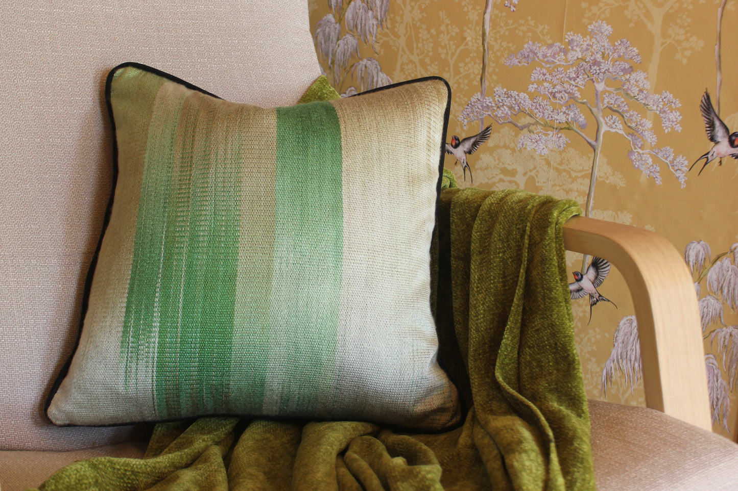 A green ombre silk cushion with black piping rests on a beige armchair, paired with a soft green throw.