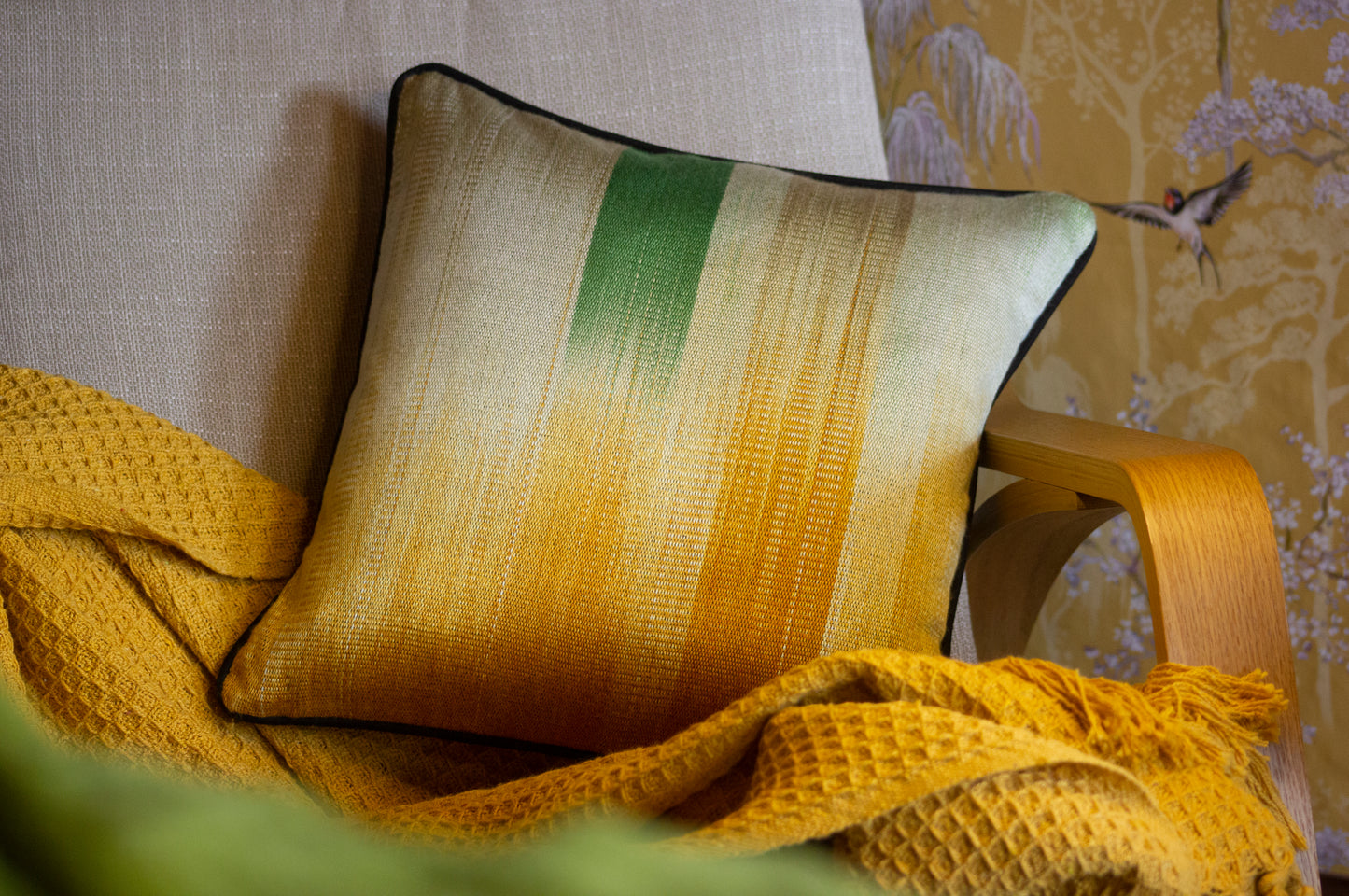 A silk cushion with a gradient of green and ochre hues rests on a beige chair with a textured yellow throw.