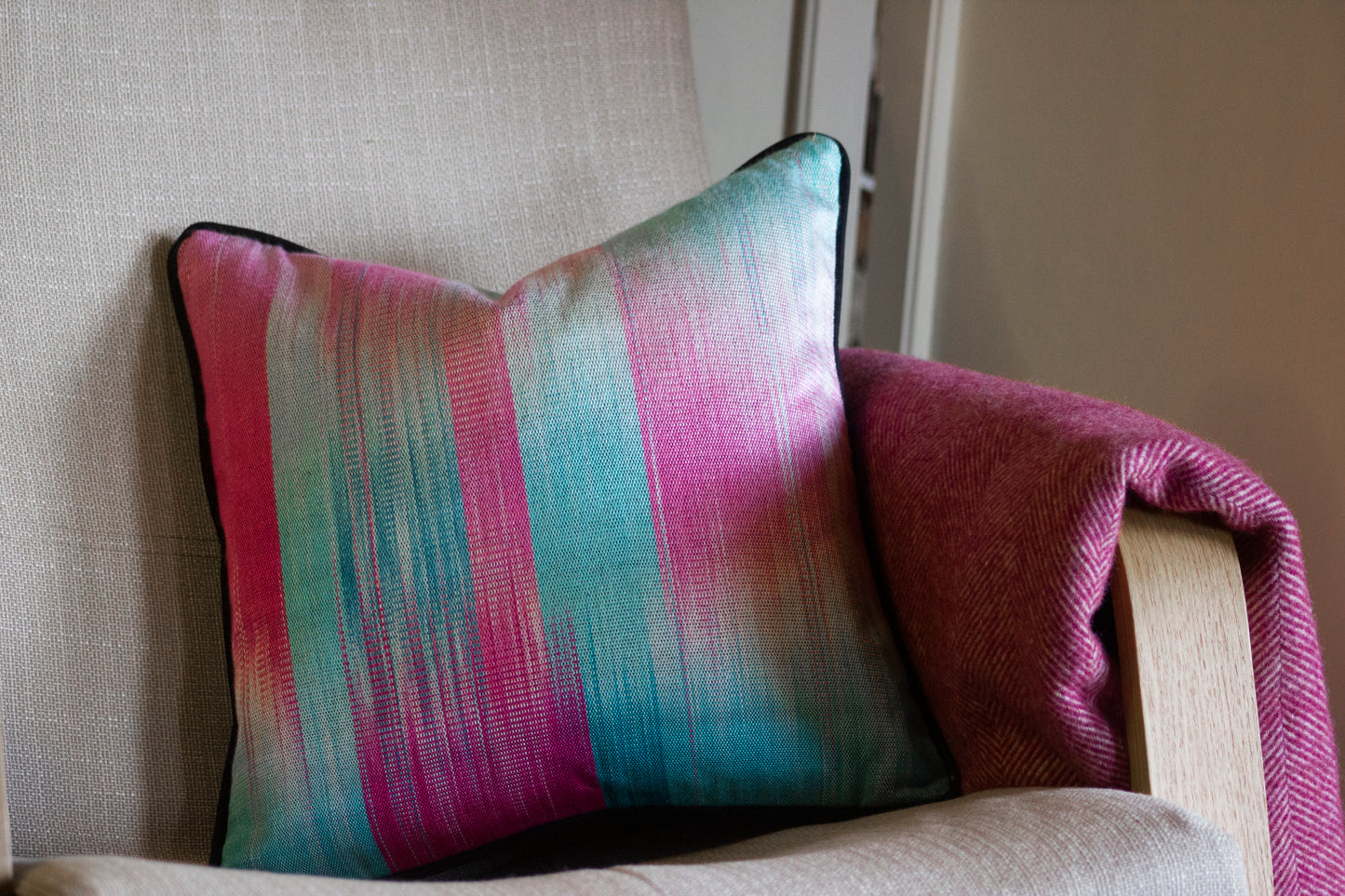 Pink and blue silk cushion with black edging on a beige chair, paired with a pink herringbone throw.