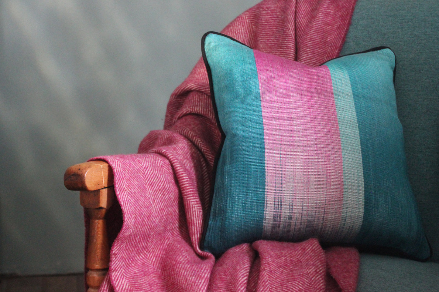 A teal and pink silk cushion on a dark turquoise chair, paired with a soft pink herringbone blanket over a wooden armrest