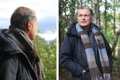 Herringbone striped alpaca scarf in blue, beige and brown. Male model wearing the scarf standing in wood. 