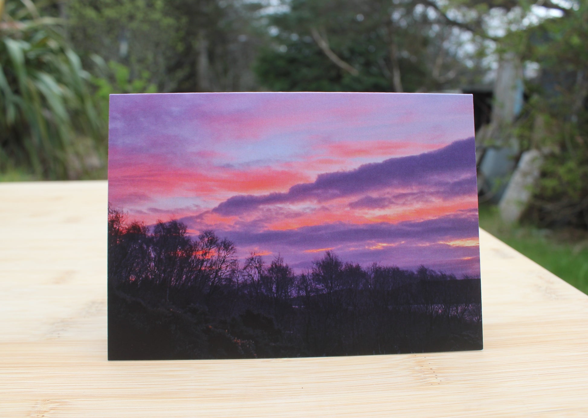 Scottish Highlands card of an intense Pink and purple evening sunset with a dark tree line in the foreground.